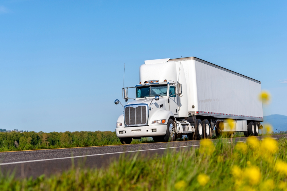 A white semi-truck driving along a scenic highway with wildflowers in the foreground.