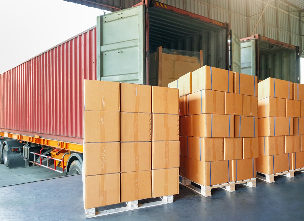 Stacked cardboard boxes on pallets beside an open red shipping container at a warehouse.